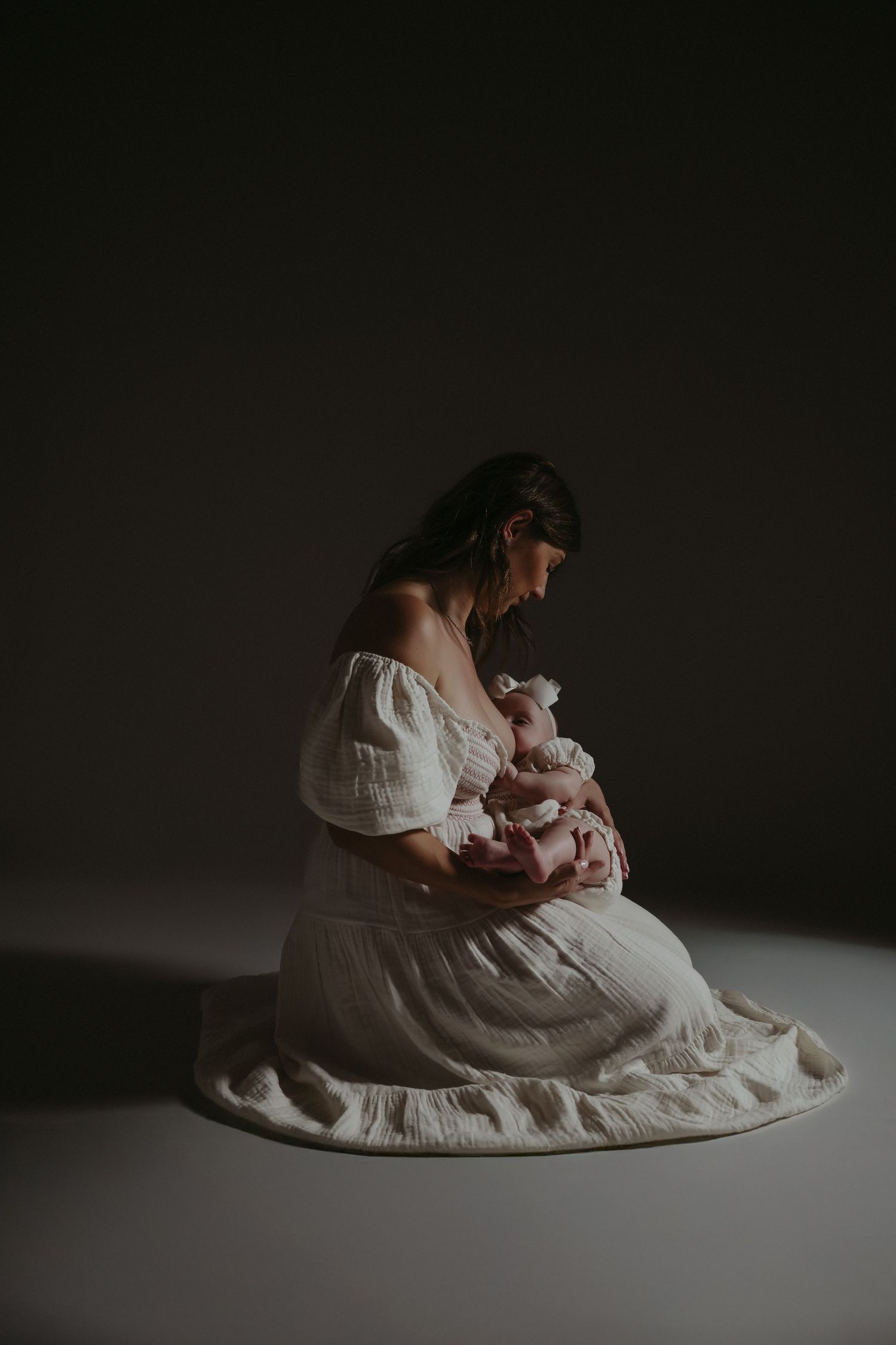 A mother in a white off-shoulder dress sits on the floor cradling her newborn baby in a softly lit studio.
