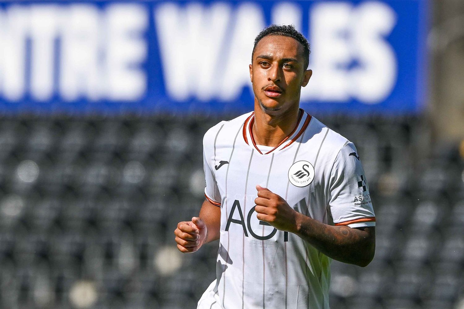 Swansea City footballer in white AO.com sponsored kit jogging on pitch with Wales signage in background.