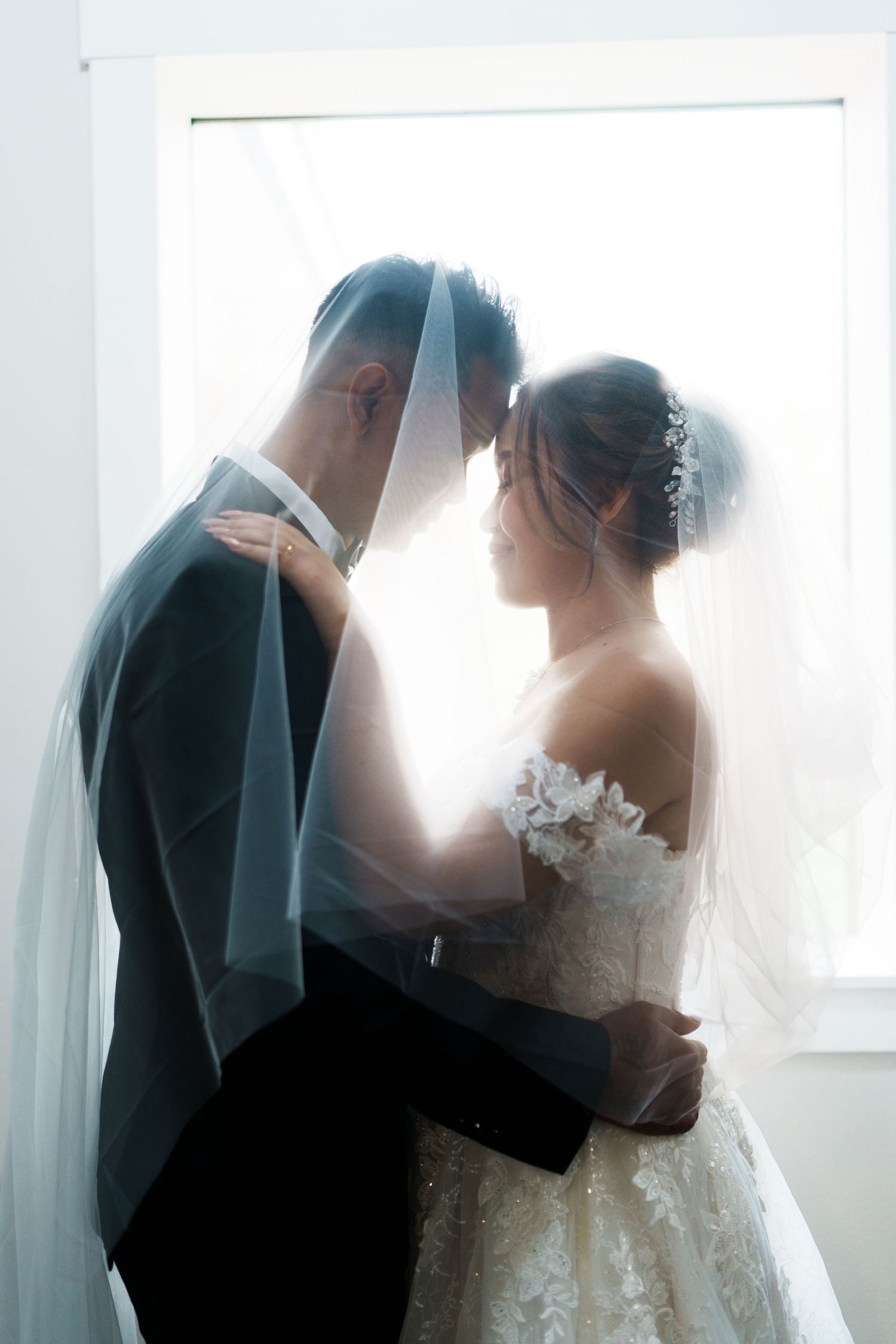 Bride and groom standing close together under a veil in soft backlit window light indoors.