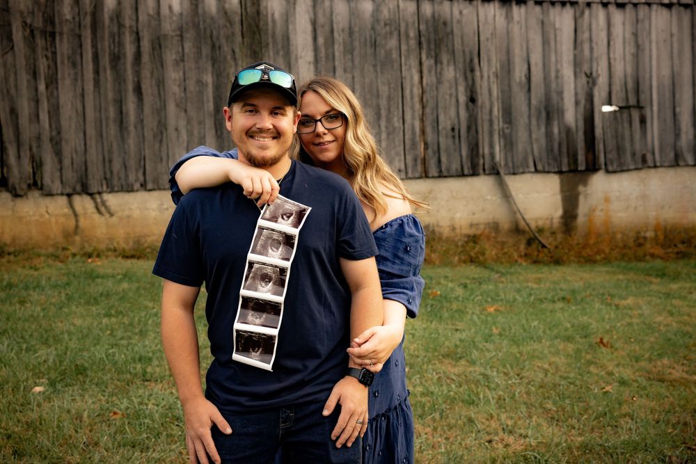 A smiling couple poses outdoors holding an ultrasound strip in front of a weathered wooden barn.