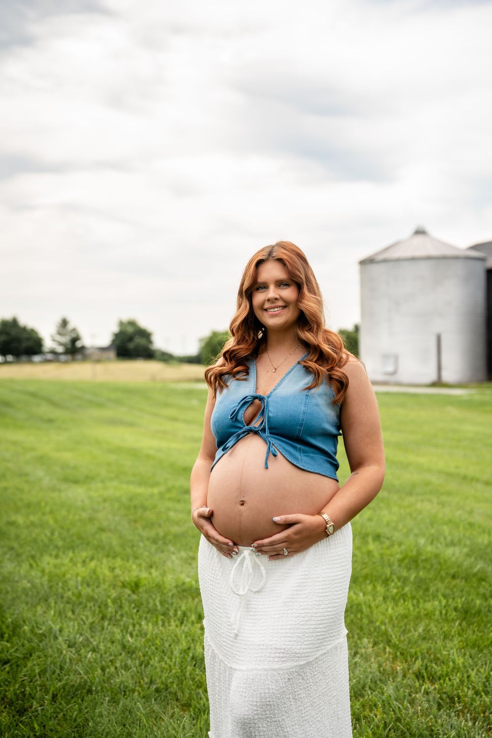Smiling pregnant woman with red hair cradling baby bump in blue crop top and white skirt outdoors.