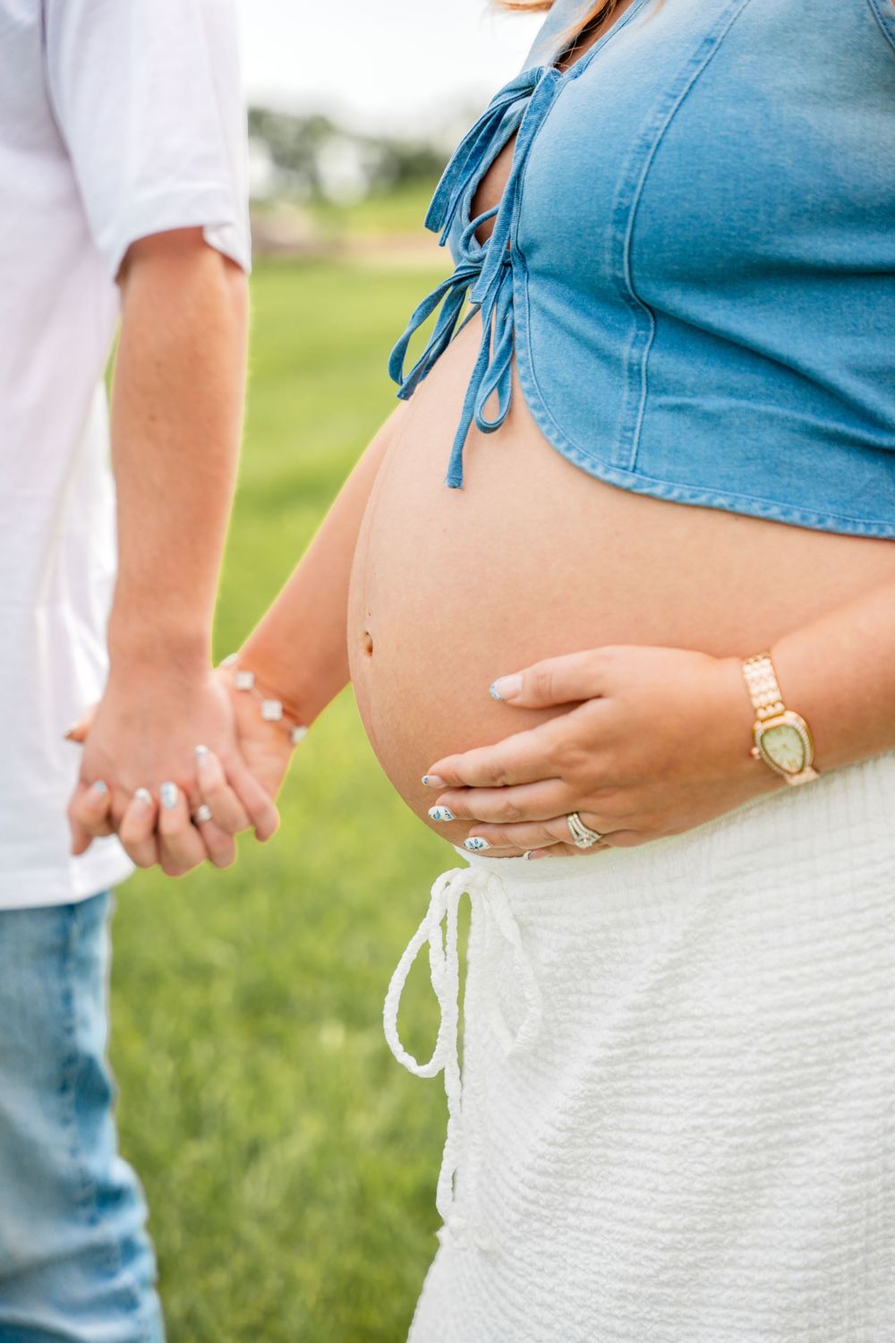 Close-up of pregnant woman in blue crop top holding baby bump while holding partner's hand outdoors.