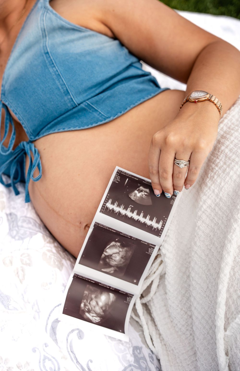 Pregnant woman in blue crop top holding ultrasound photos against her baby bump while reclining outdoors.