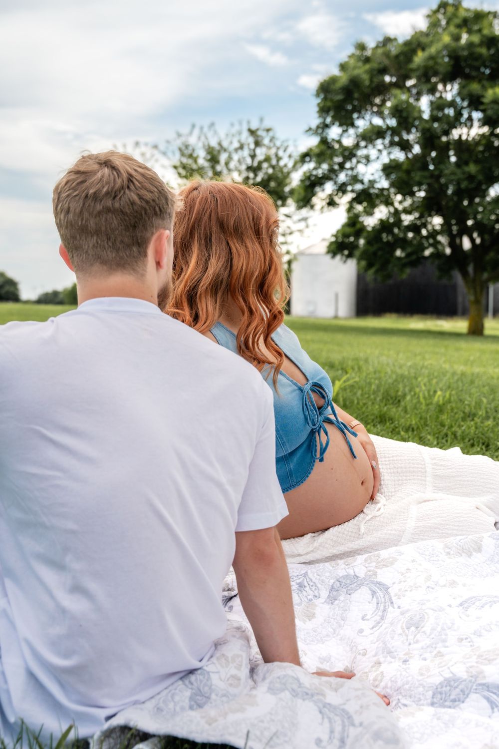 Couple sitting on blanket in green field, man with arm around pregnant woman holding her baby bump.