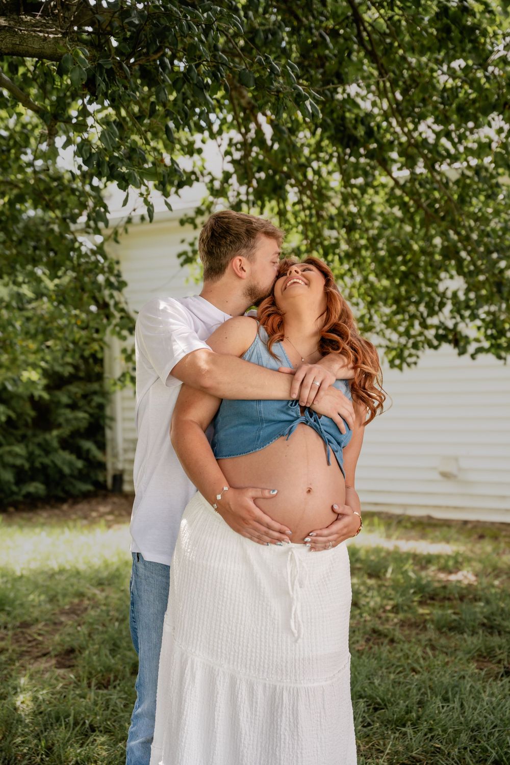Couple embracing outdoors under green tree, man kissing pregnant woman's cheek while holding her baby bump.