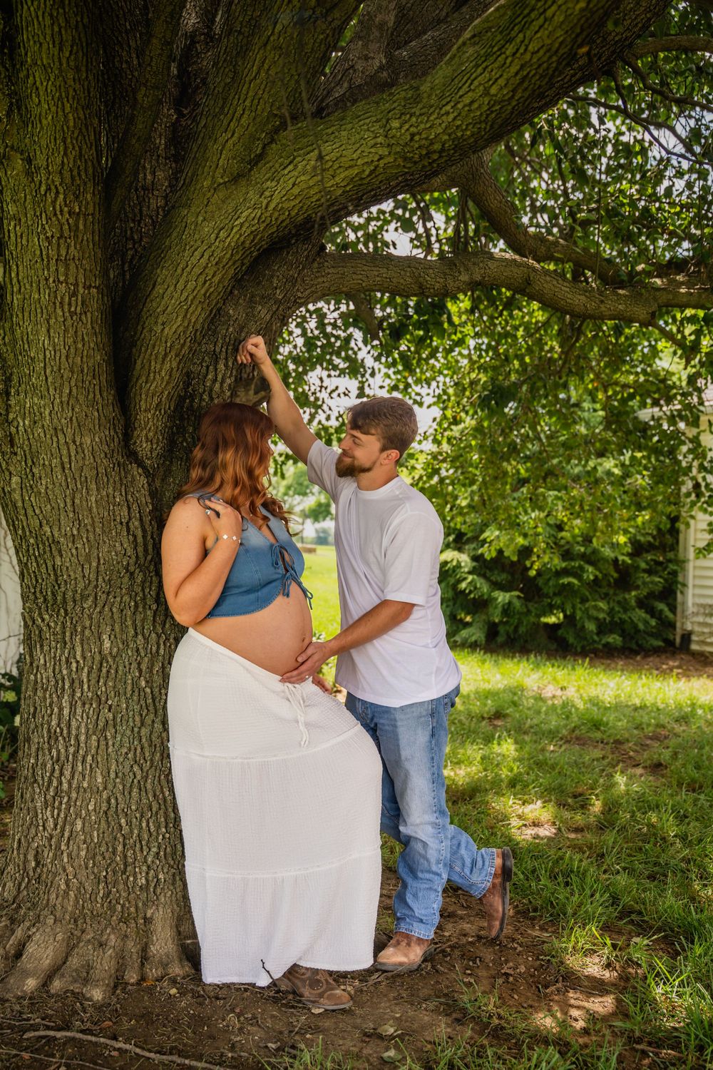 Couple sharing tender moment under large oak tree, man touching pregnant woman's baby bump outdoors.