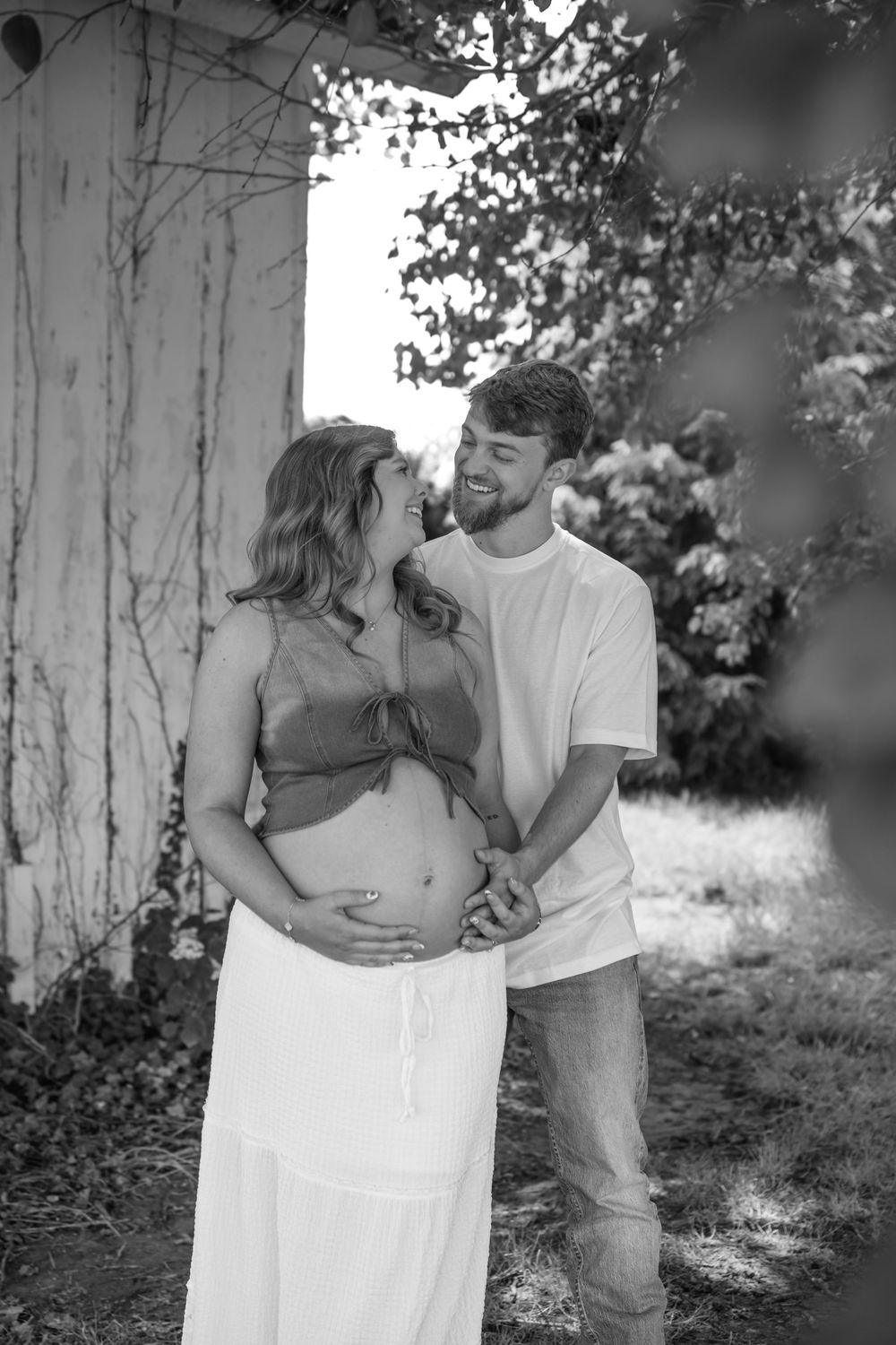 Black and white photo of smiling couple, pregnant woman leaning into partner outdoors near trees.