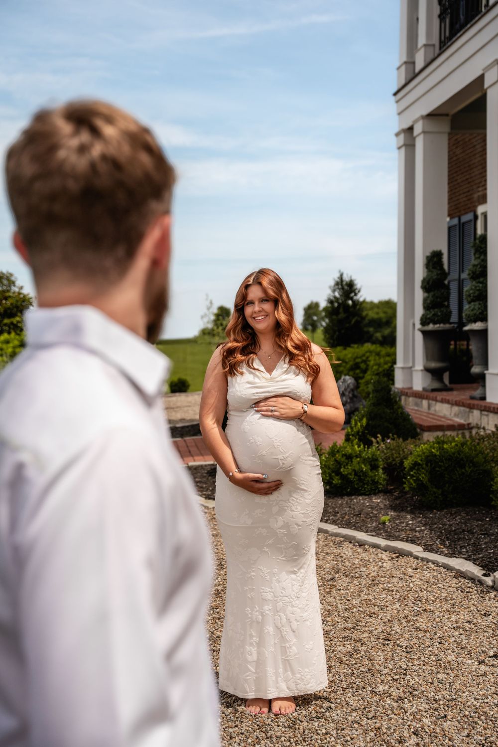 Smiling pregnant woman in white lace gown cradling baby bump outside elegant estate on sunny day.