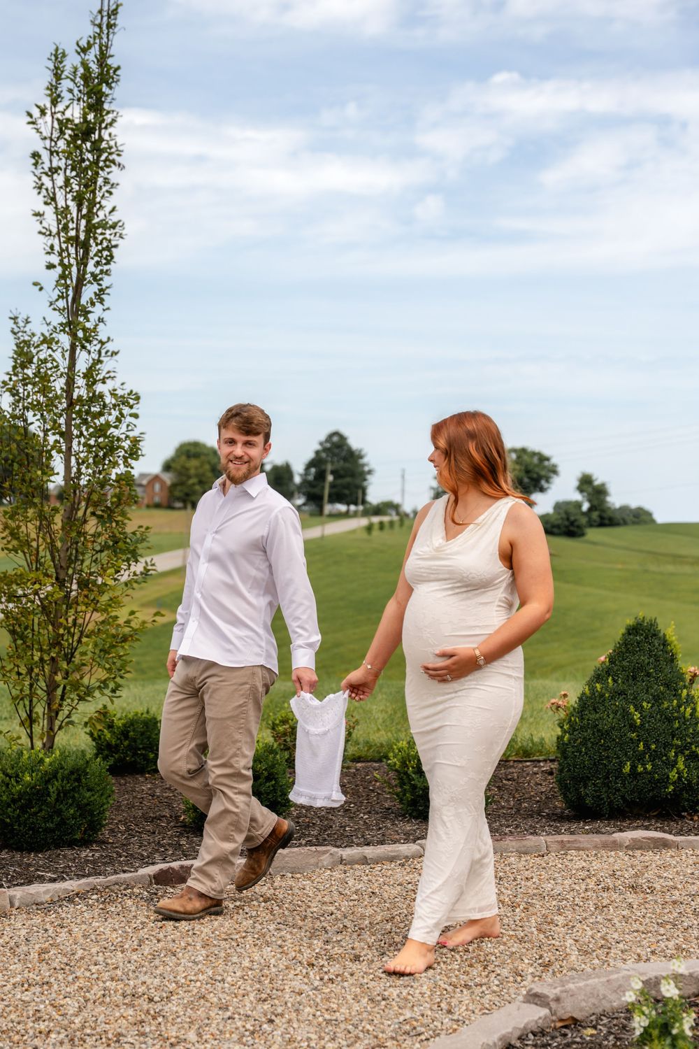Couple walking on gravel path, man holding tiny baby outfit while pregnant woman touches her bump.