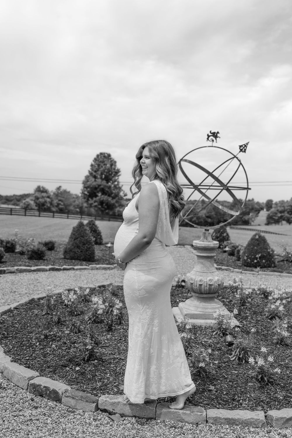 Black and white portrait of pregnant woman in white gown beside armillary sphere in garden setting.