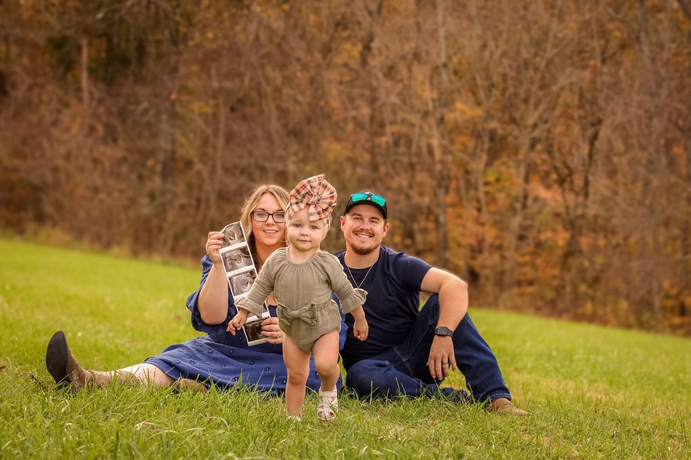 A young family with a toddler sits on green grass in front of colorful autumn foliage.