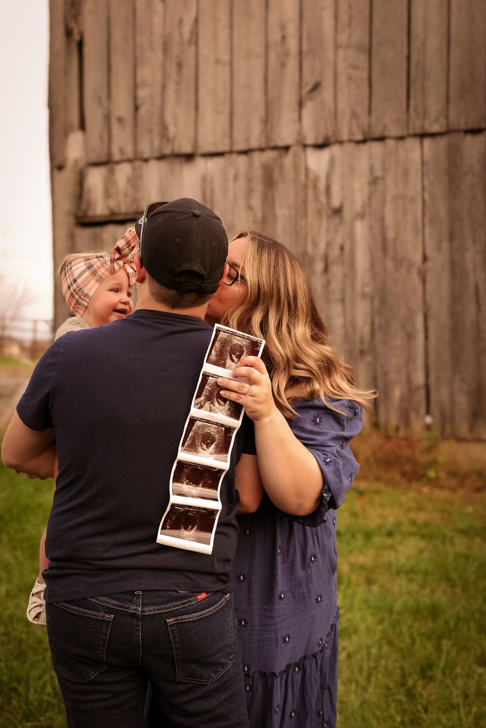 A couple kisses while holding ultrasound photos with a toddler beside them near a rustic barn.