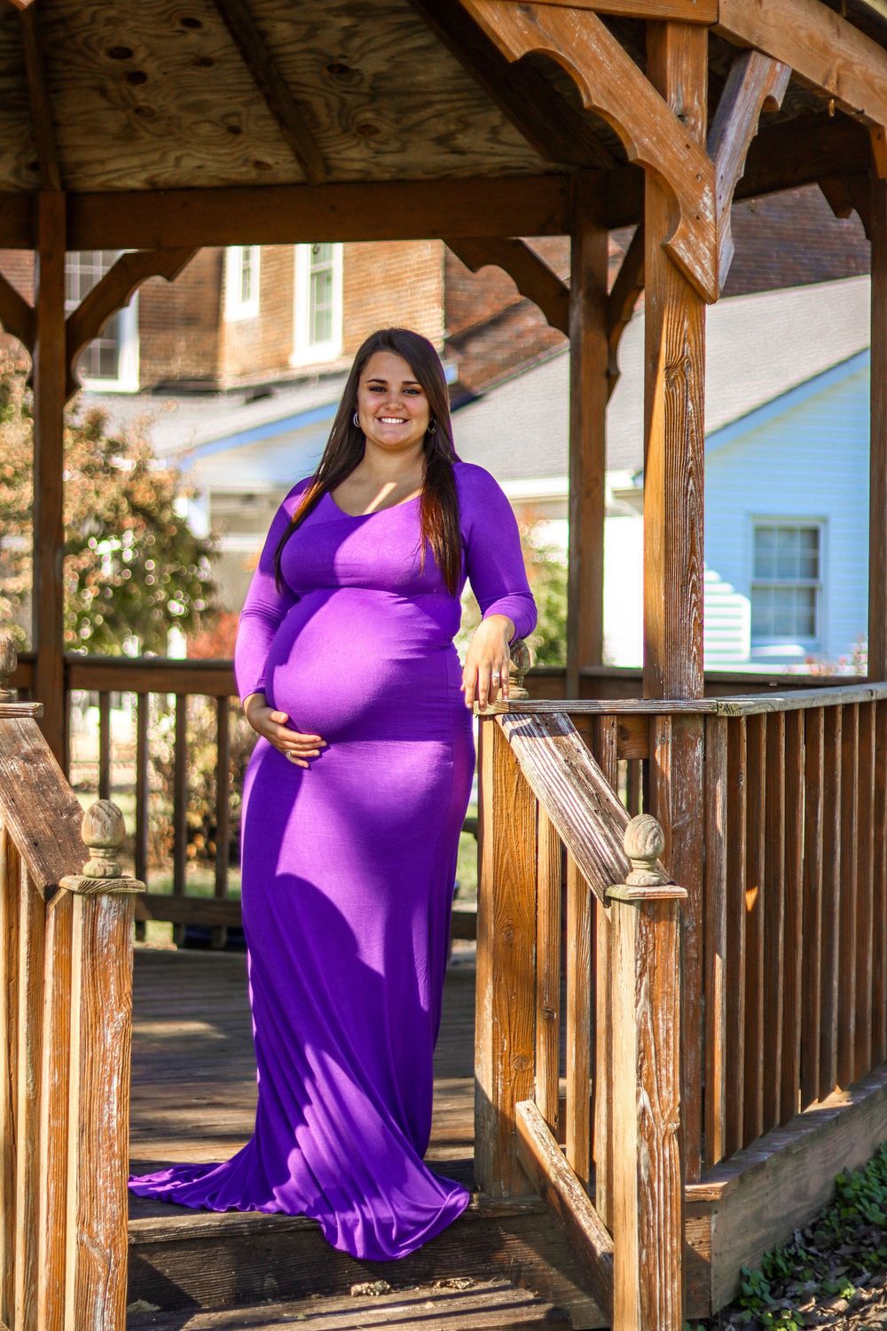 Smiling pregnant woman in purple gown cradling baby bump while standing in wooden gazebo outdoors.