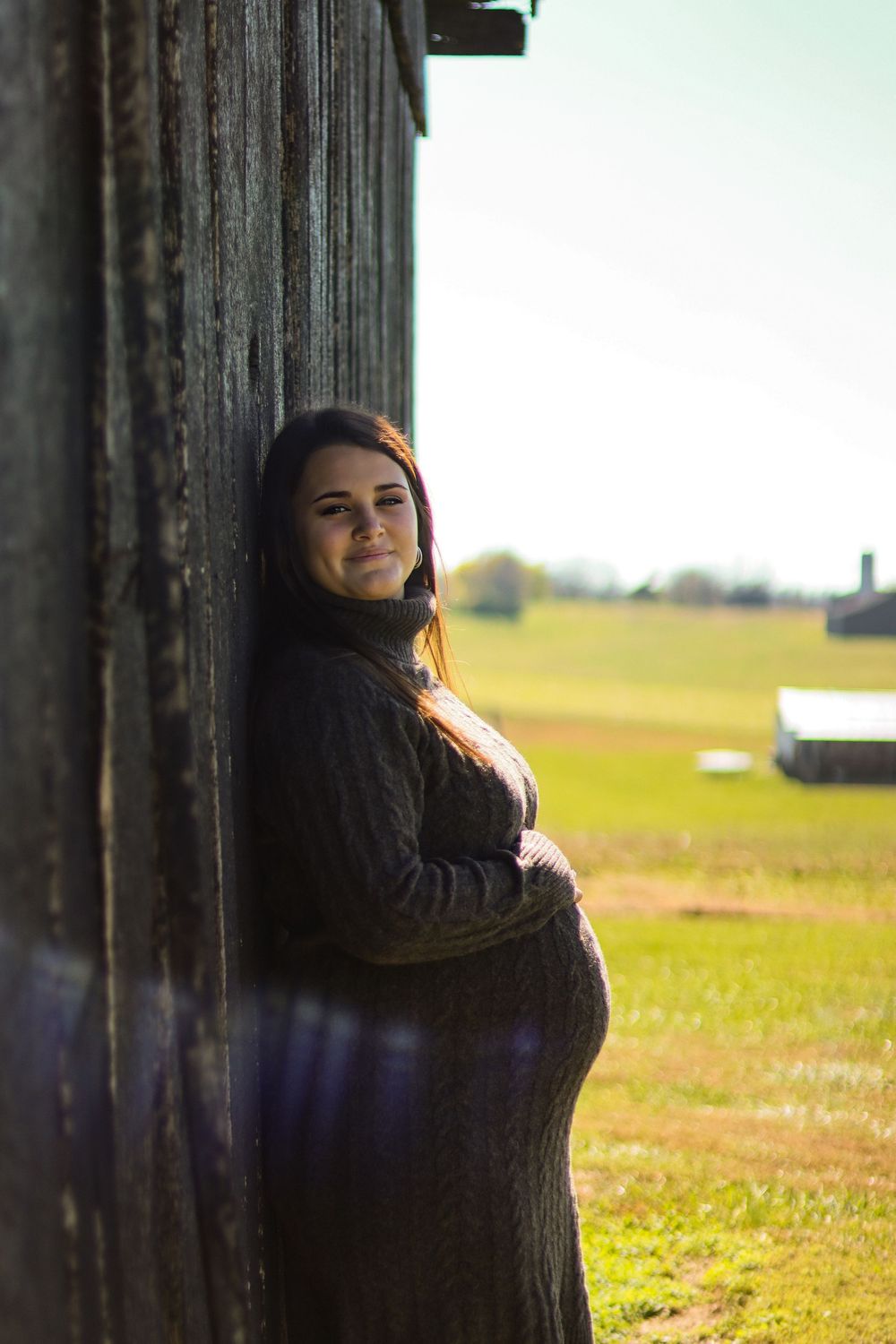 Pregnant woman in dark knit dress leaning against weathered barn wall in sunlit rural field.