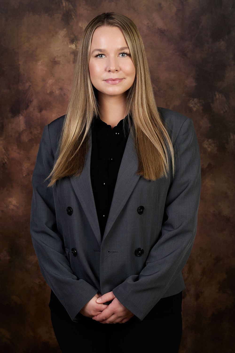 Professional woman in gray blazer and black top poses confidently against a brown studio backdrop.