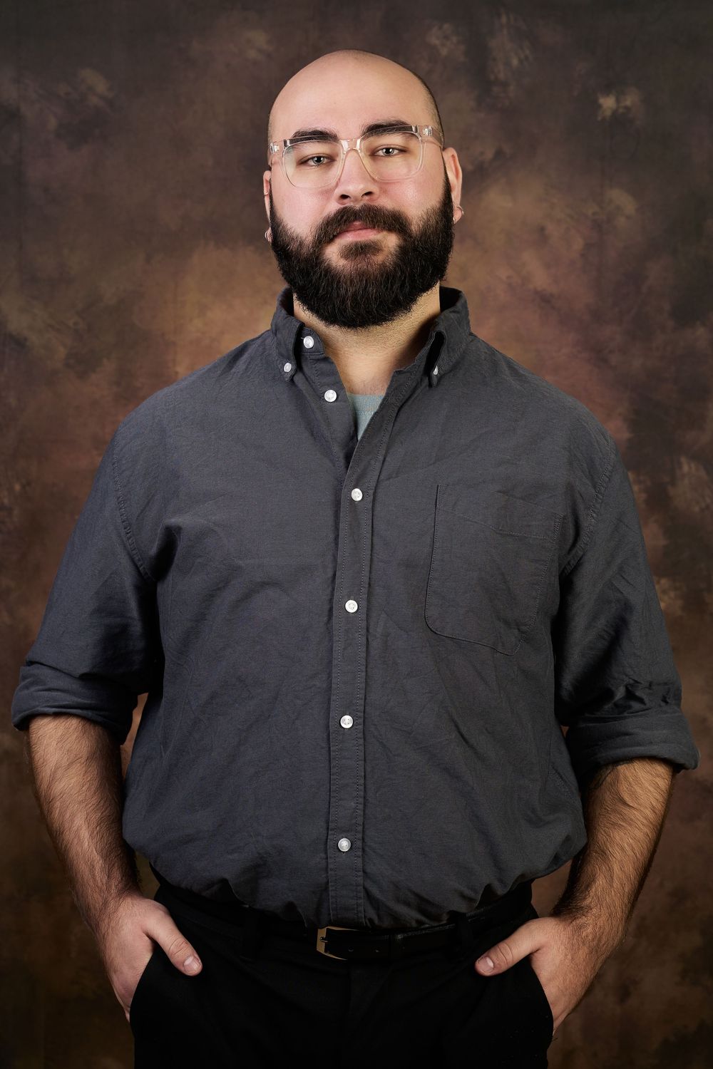 Bald bearded man wearing glasses and a gray button-up shirt poses with hands in pockets against a brown backdrop.