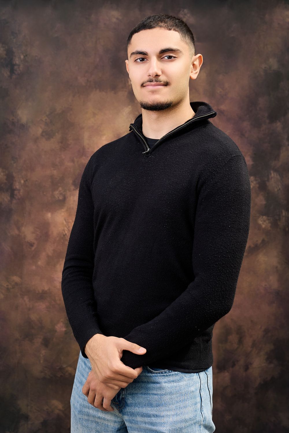 Young man in black zip-neck sweater and jeans posing confidently against a brown studio backdrop.