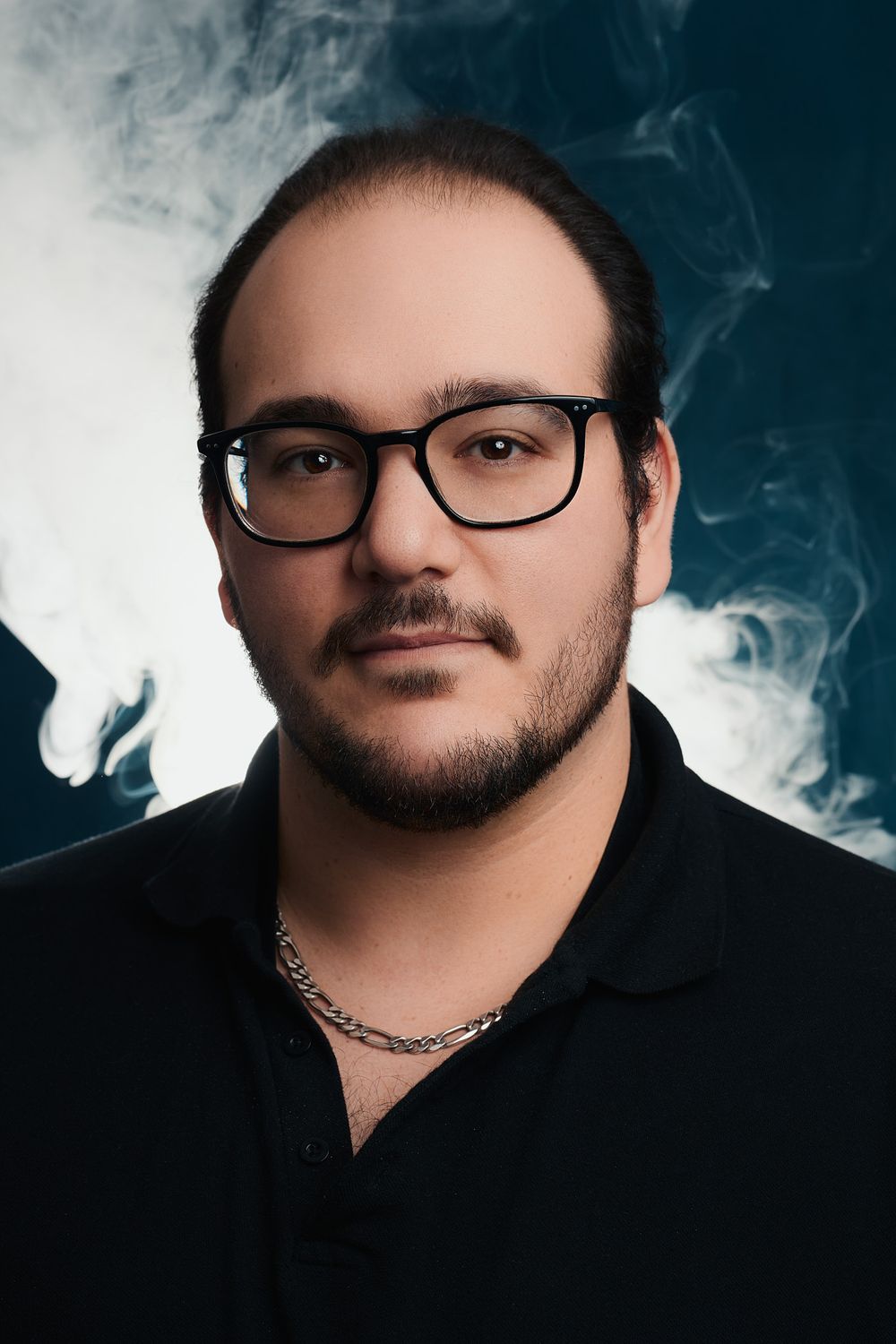 A young man with glasses, beard, and chain necklace poses against a dramatic smoky dark blue background.