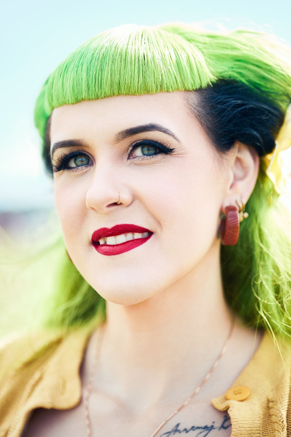 Young woman with vibrant green hair, red lips, nose ring, and hoop earrings smiling outdoors.