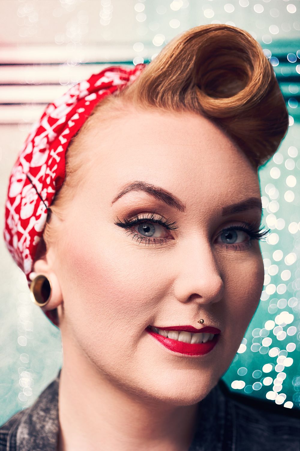 A smiling woman with retro pin-up style, red bandana headband, victory rolls, red lipstick, and cat-eye makeup.