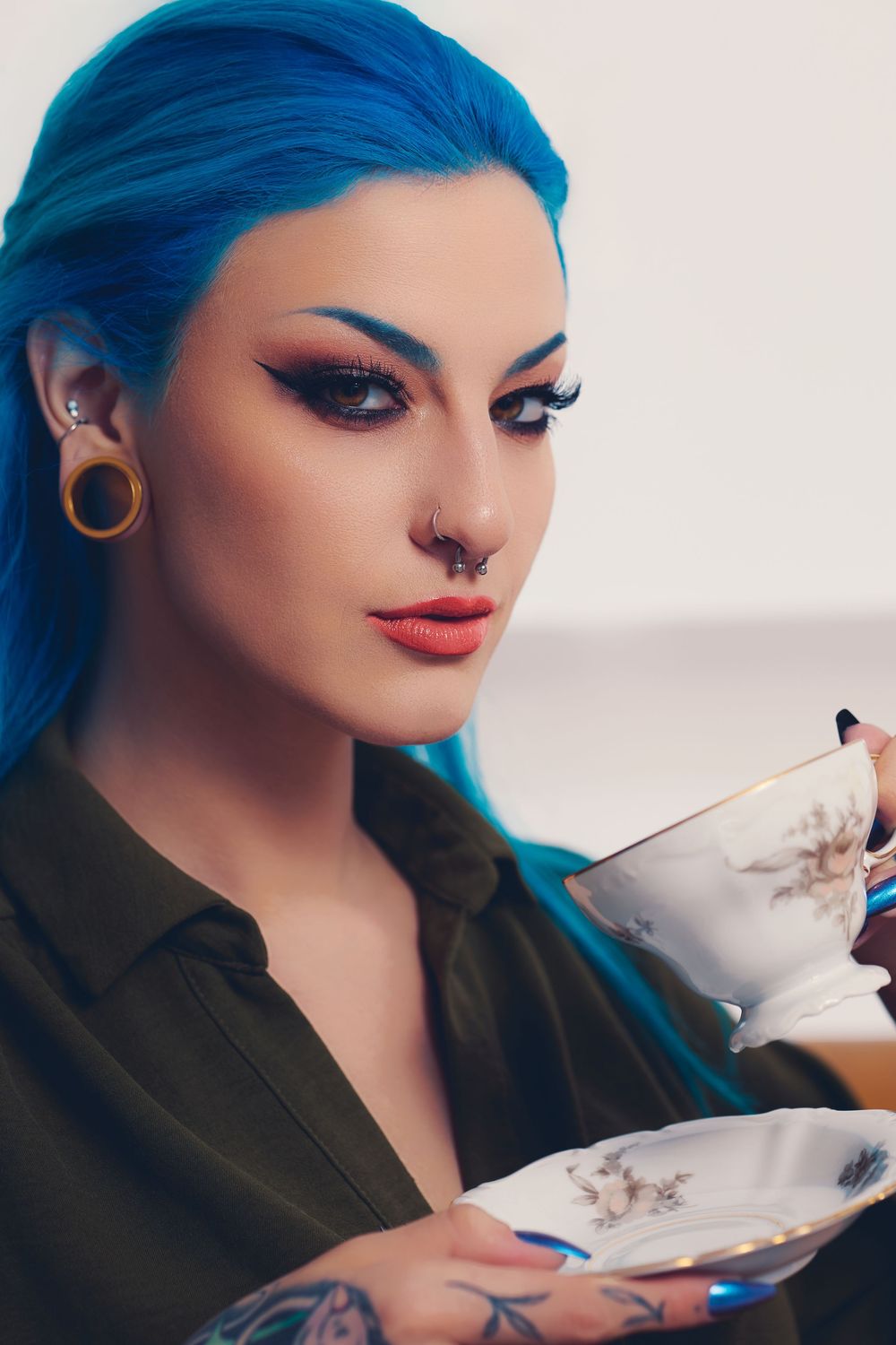 Woman with blue hair, tattoos, and piercings holding a vintage teacup and saucer with dramatic eye makeup.