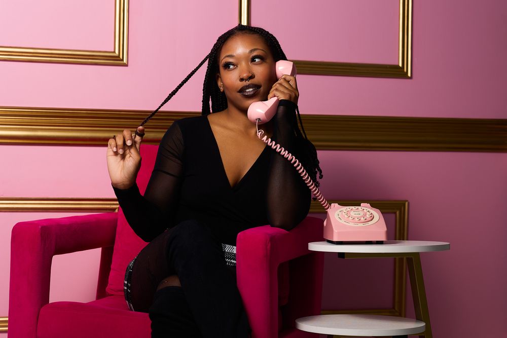 Woman in black outfit sitting in pink chair, holding vintage pink rotary phone in elegant pink room.