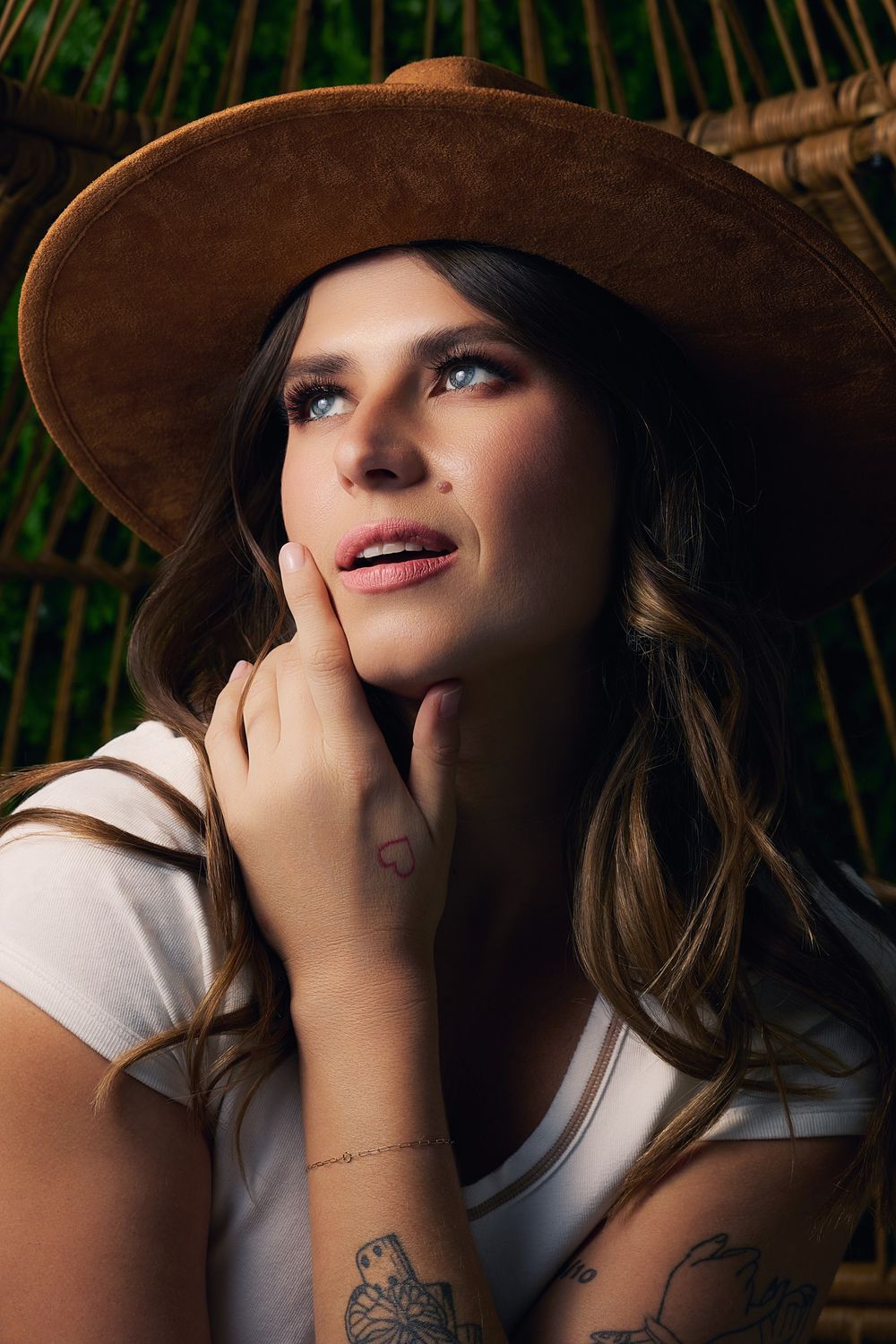 Young woman in brown wide-brim hat with tattoos gazes upward thoughtfully, hand on cheek, against bamboo background.