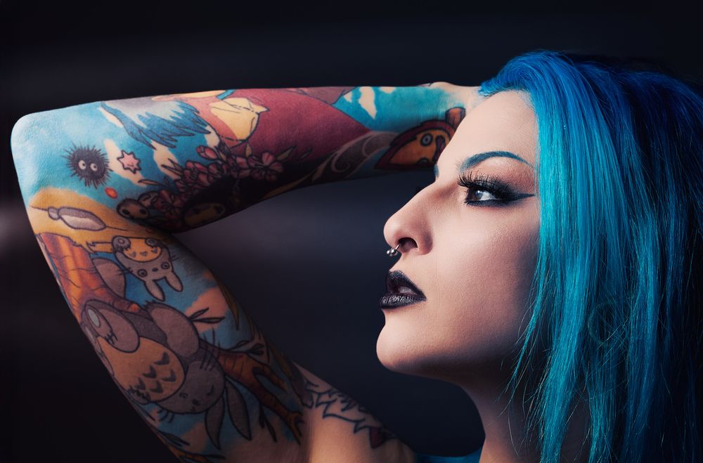 Woman with blue hair, dark makeup, and colorful tattoo sleeve posing against a dark background.