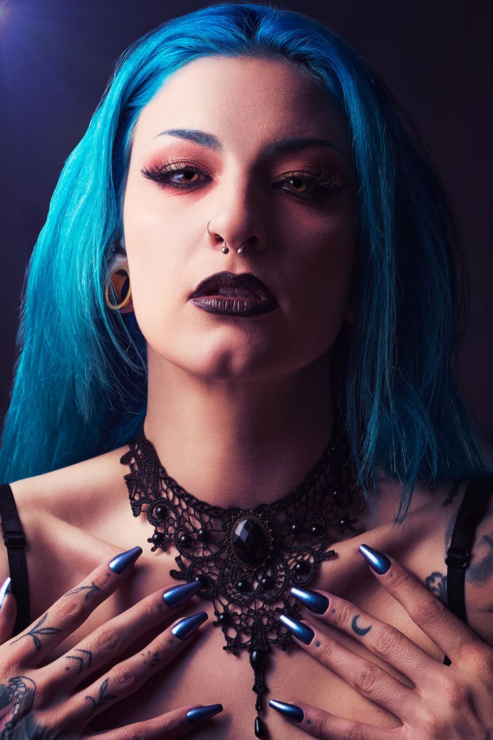 Woman with blue hair, dark lipstick, lace choker, chrome nails, and tattoos poses dramatically against dark background.