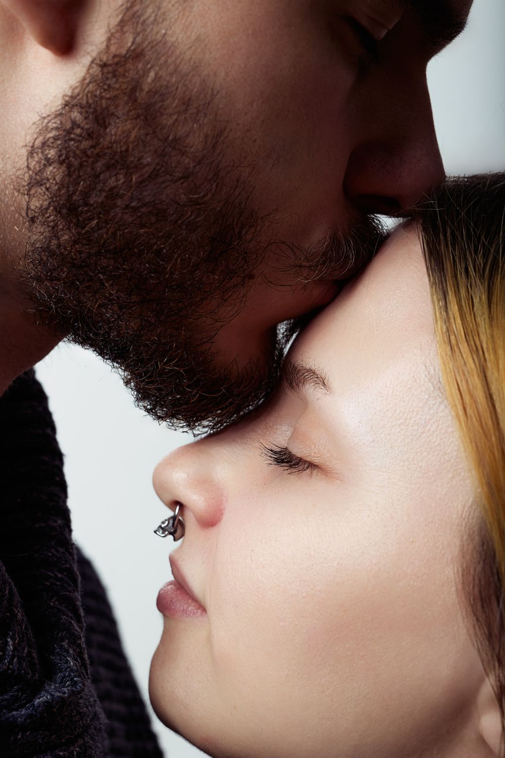 A bearded man gently kisses a young woman with a nose piercing on the forehead in a tender romantic moment.