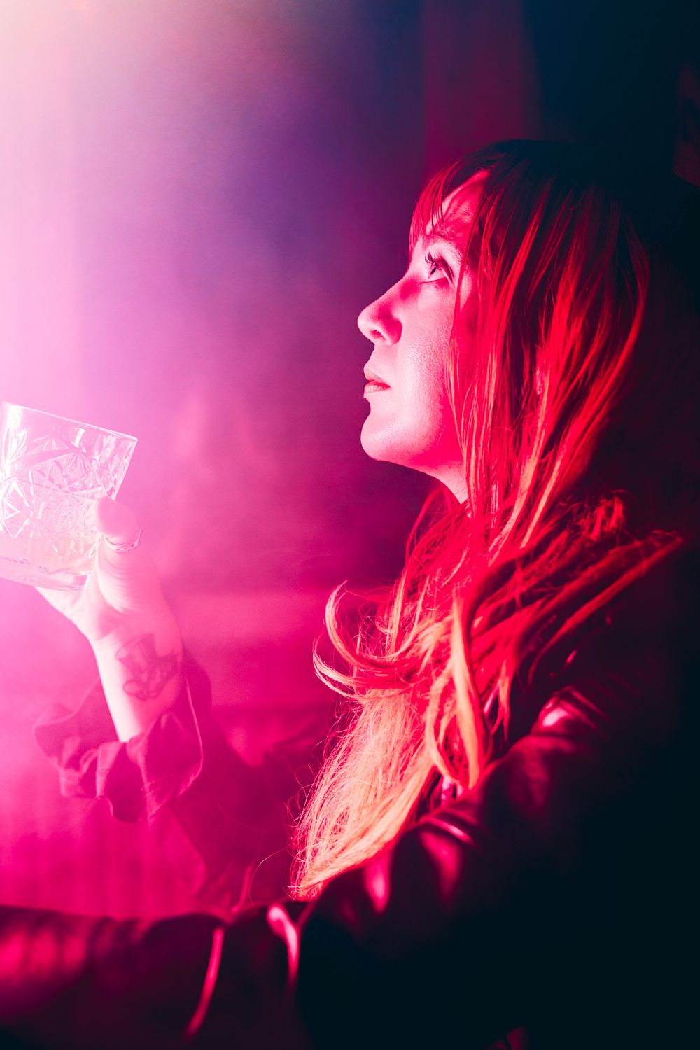 Woman in leather jacket holding a crystal glass under dramatic pink and red nightclub lighting.