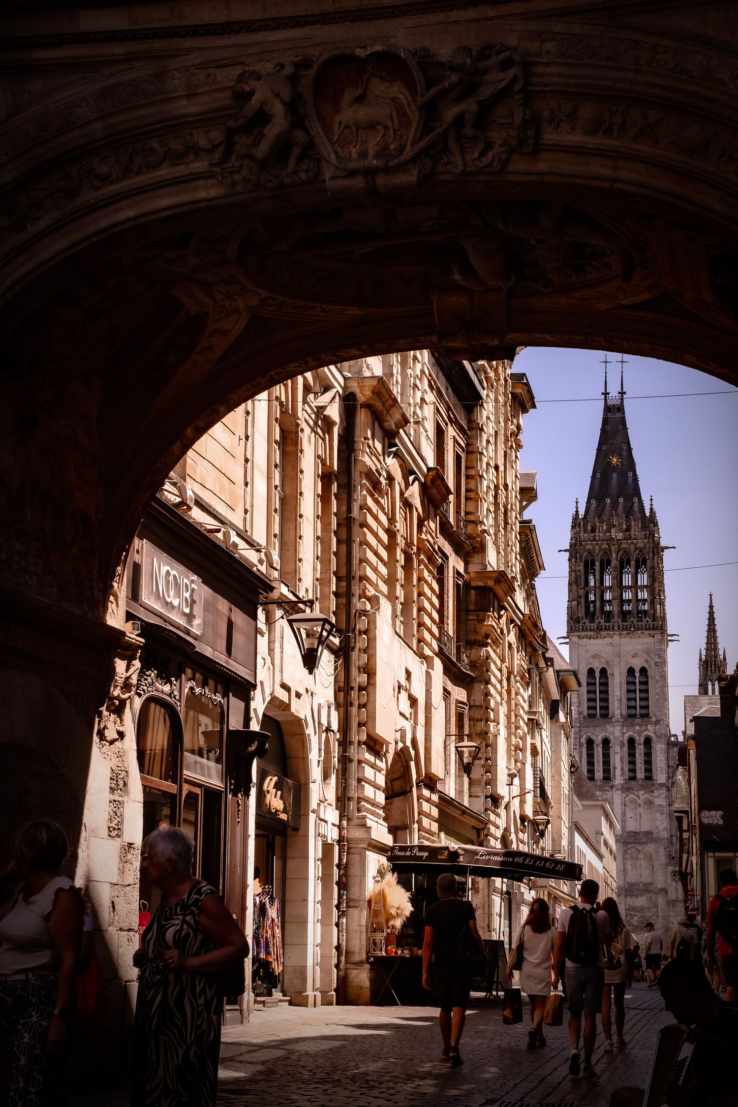 Historic European street viewed through ornate arch, with Gothic church tower rising above old stone buildings and pedestrians.