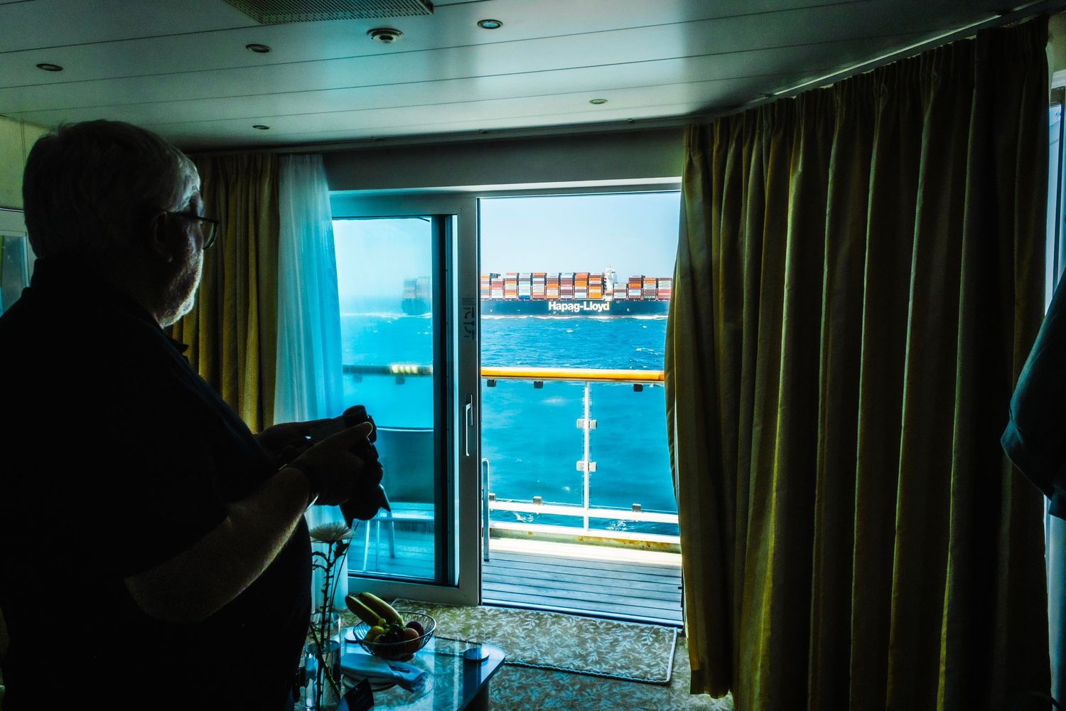 Person standing in cruise ship cabin looking out window at large cargo ship on blue ocean water.