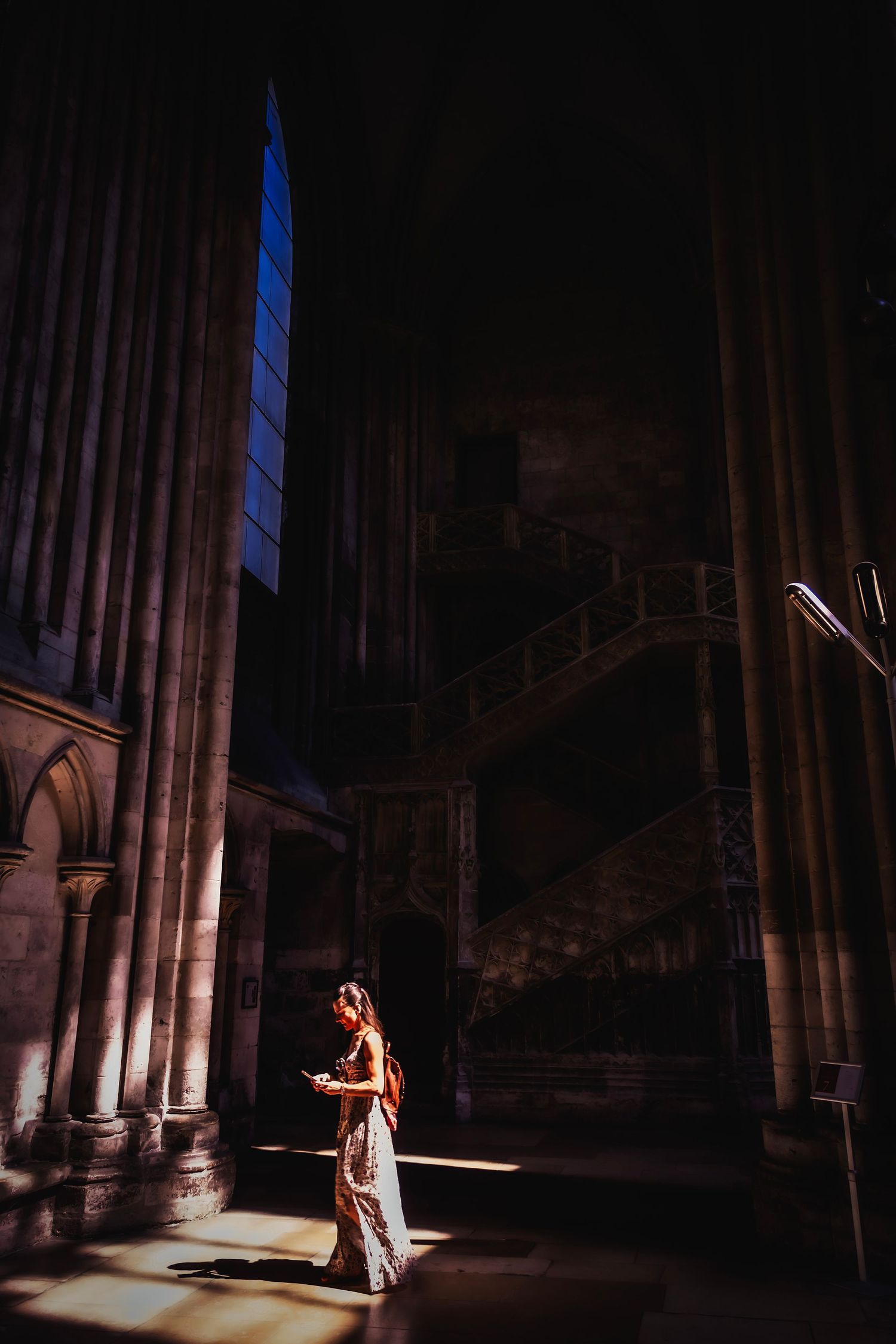 Woman in elegant gown standing in dramatic cathedral light with towering Gothic columns and stained glass window.