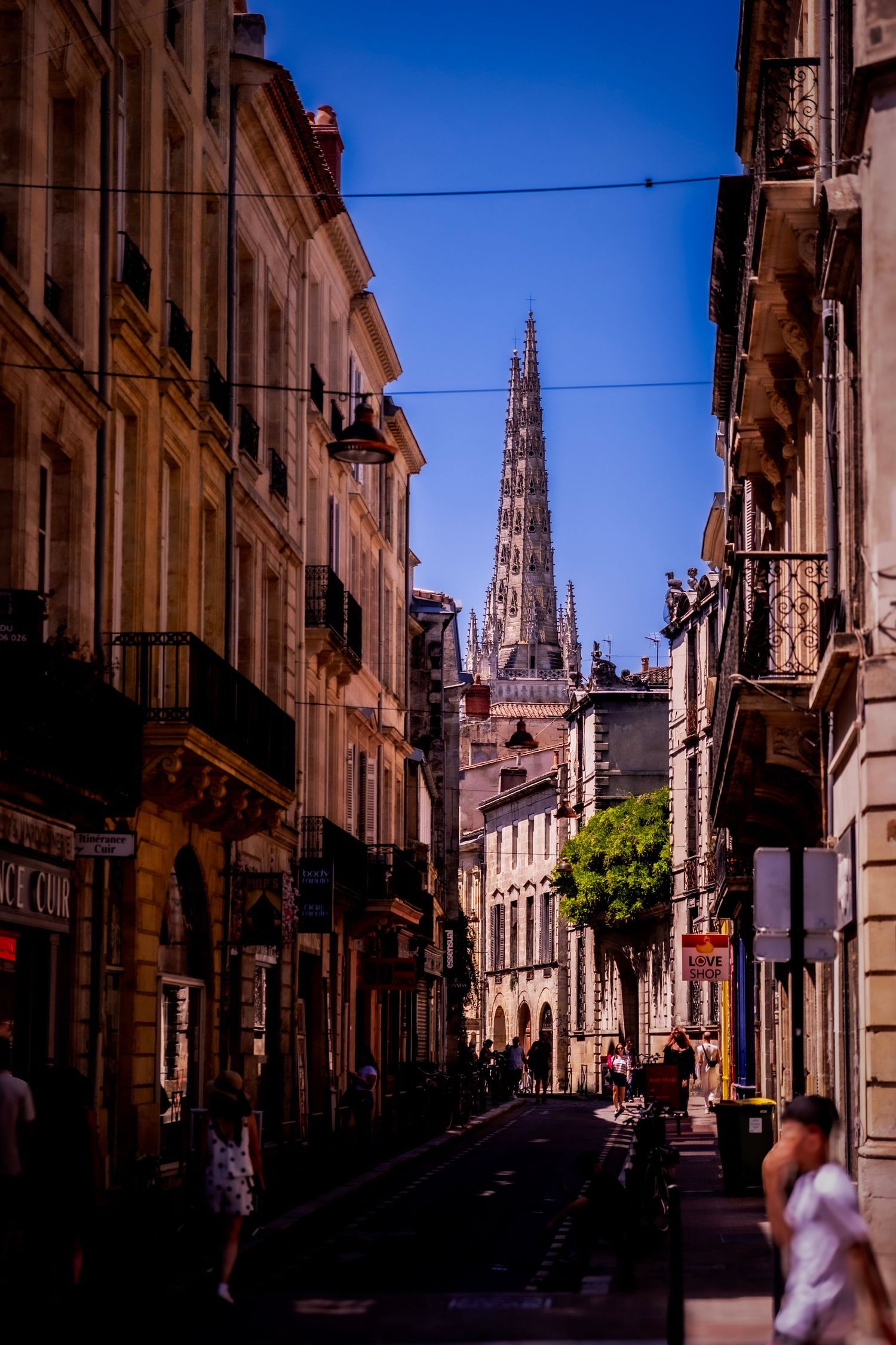 A narrow French street leads to a Gothic cathedral spire under a clear blue sky in Bordeaux, France.