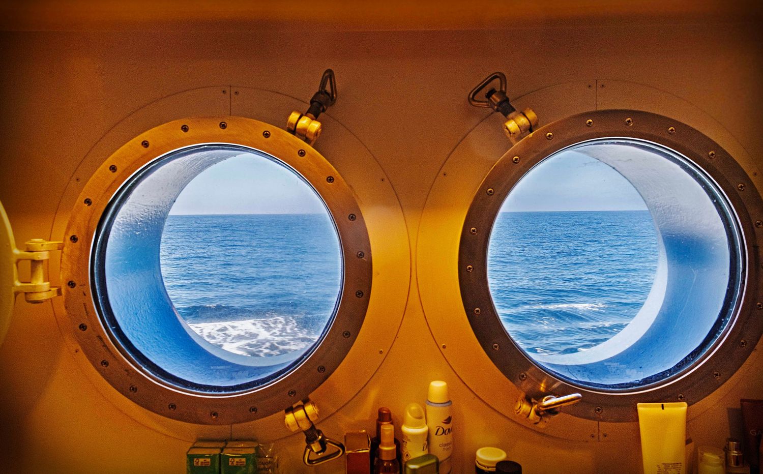 Two round brass portholes on a cruise ship overlooking the blue ocean waves from a cabin interior.