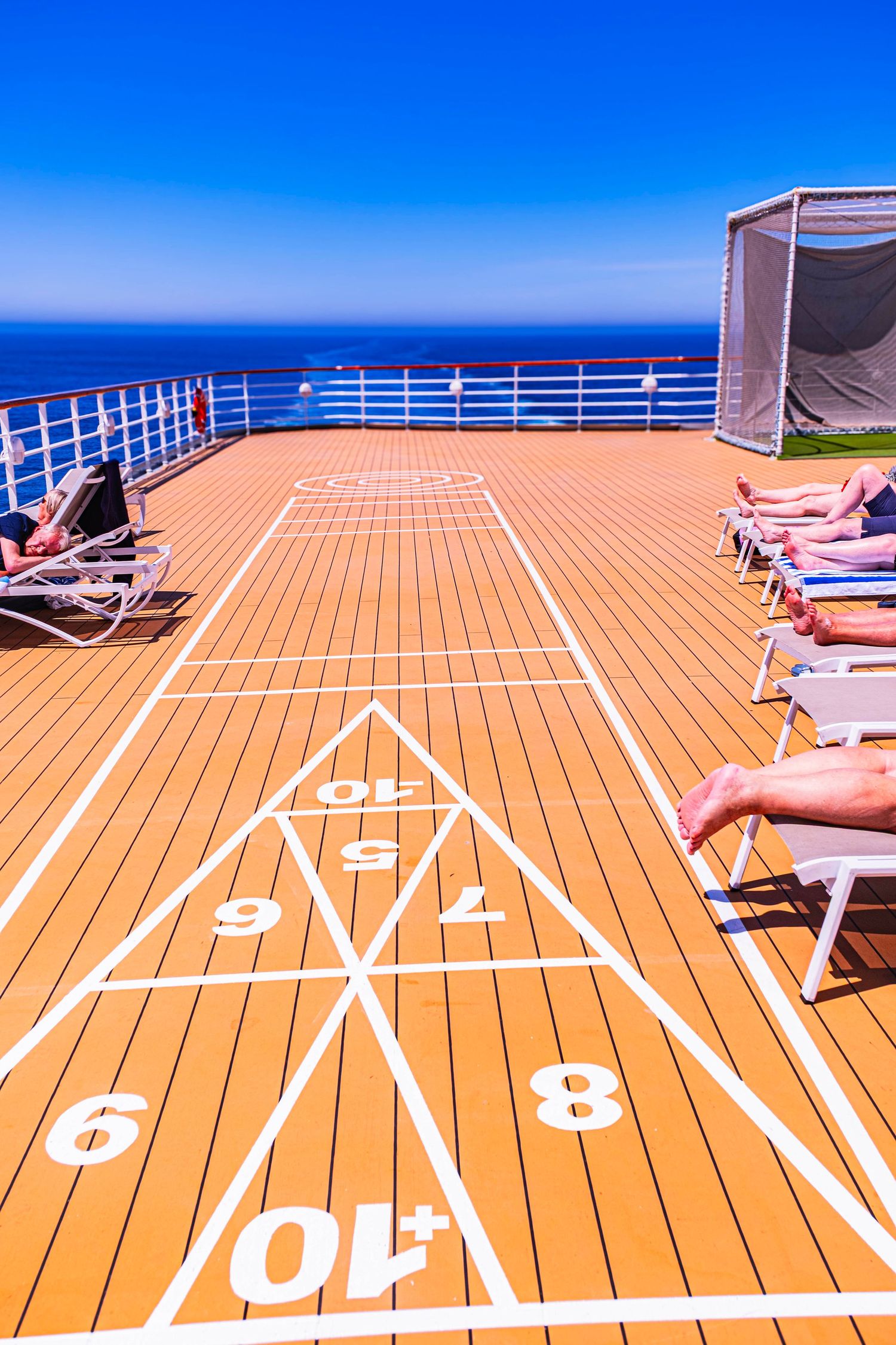 Shuffleboard court on a sunny cruise ship deck with lounge chairs and ocean views under a clear blue sky.