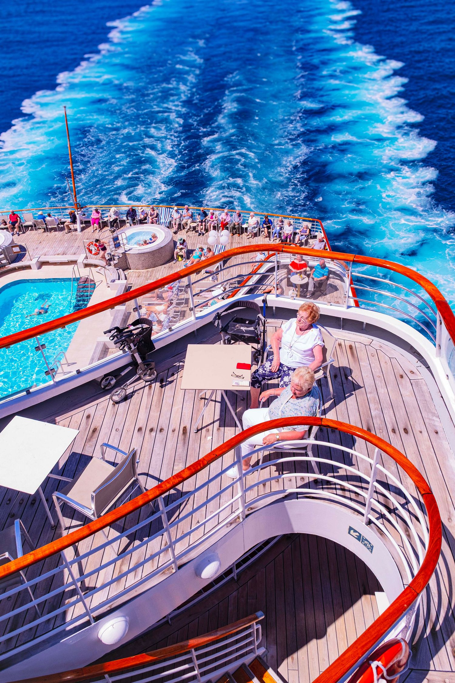 Aerial view of cruise ship deck with pool, red railings, passengers relaxing, and white wake trail in blue ocean.