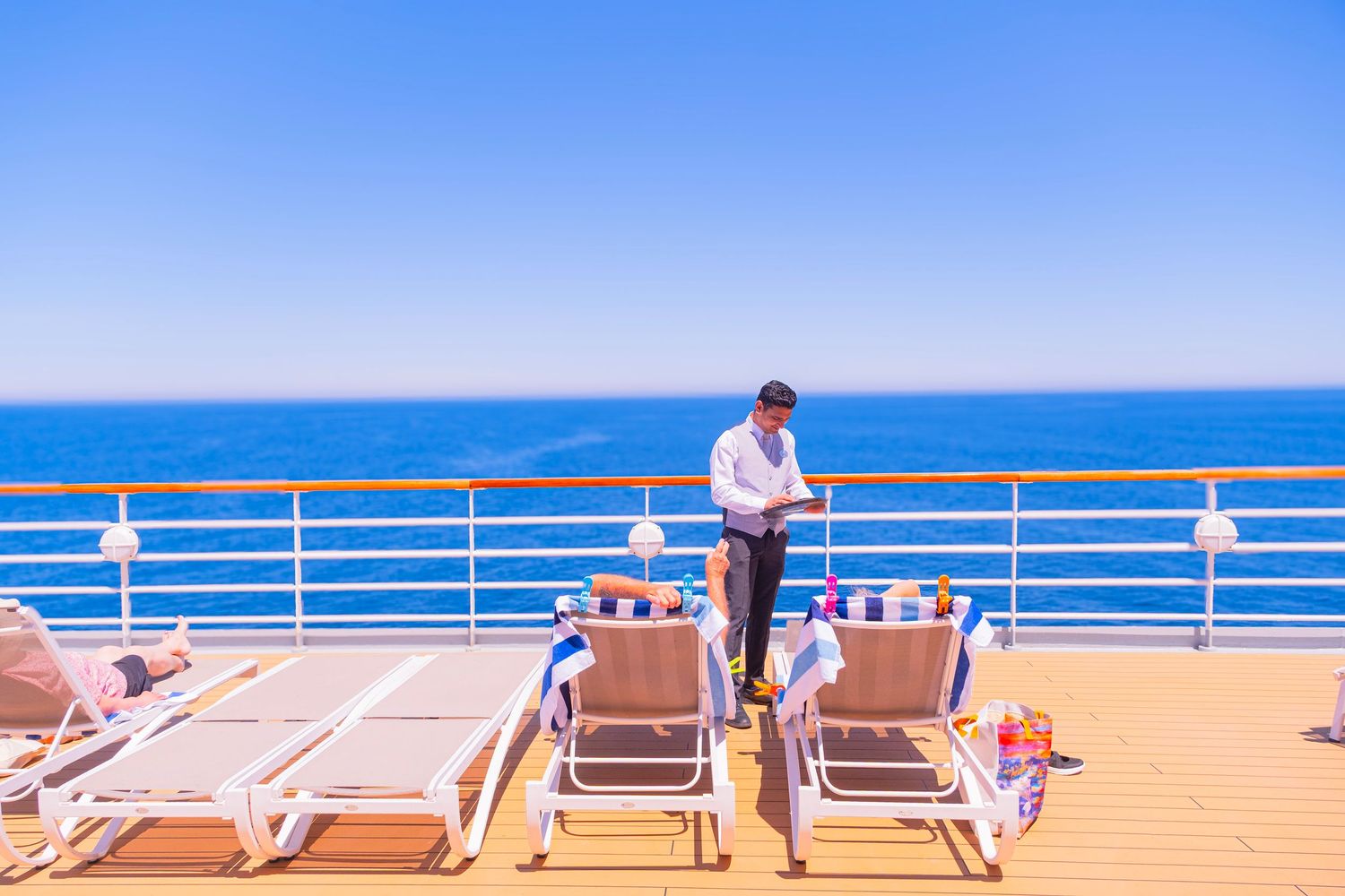 Man standing on sunny cruise ship deck with lounge chairs overlooking calm blue ocean under clear sky.