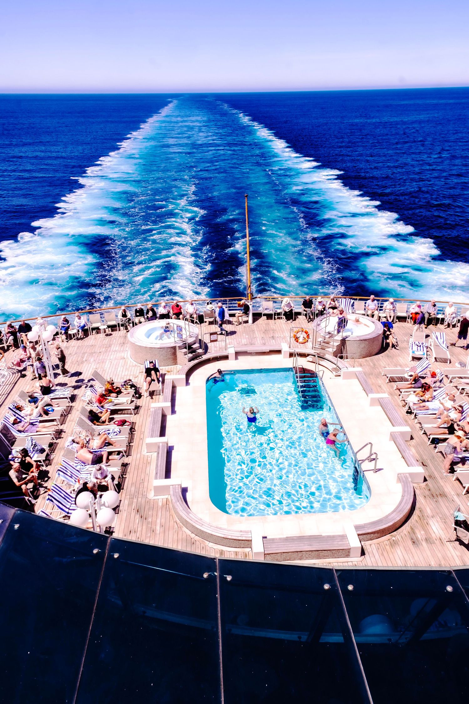 Aerial view of cruise ship pool deck with sunbathers, blue swimming pool, and white wake trail on open ocean.