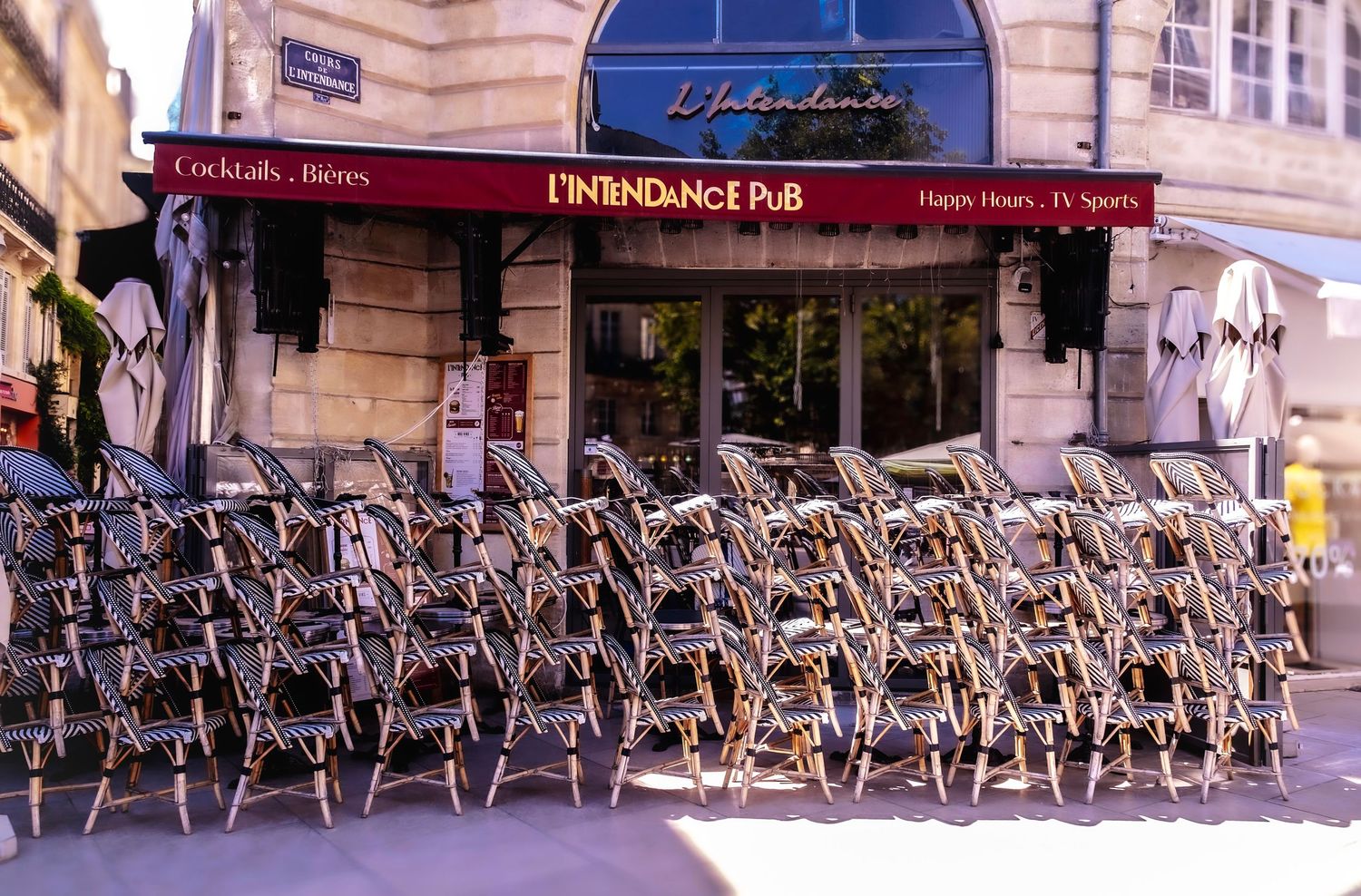 Stacked chairs outside L'Intendance Pub in Bordeaux, France, with a red awning and classic French architecture.