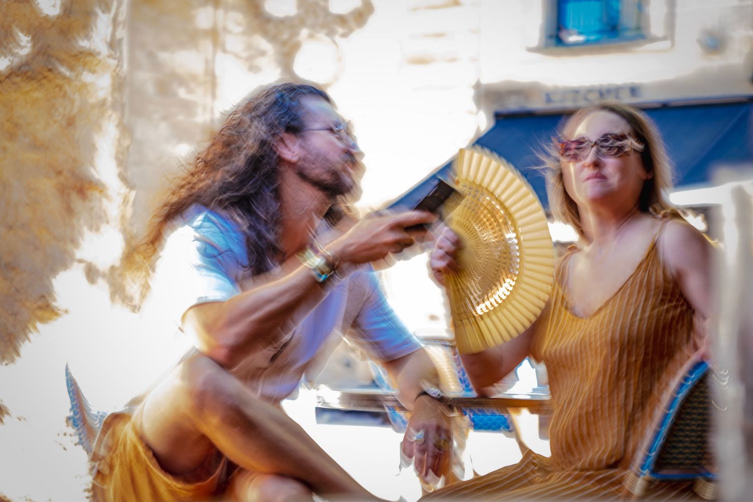 Man with long hair playing guitar for woman with sunglasses outdoors on sunny day.