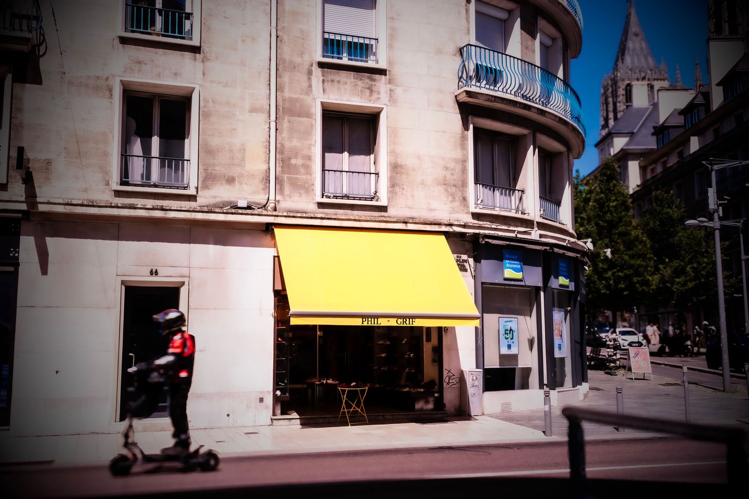 Person on electric scooter passing a French café with yellow awning on a quiet European street corner.