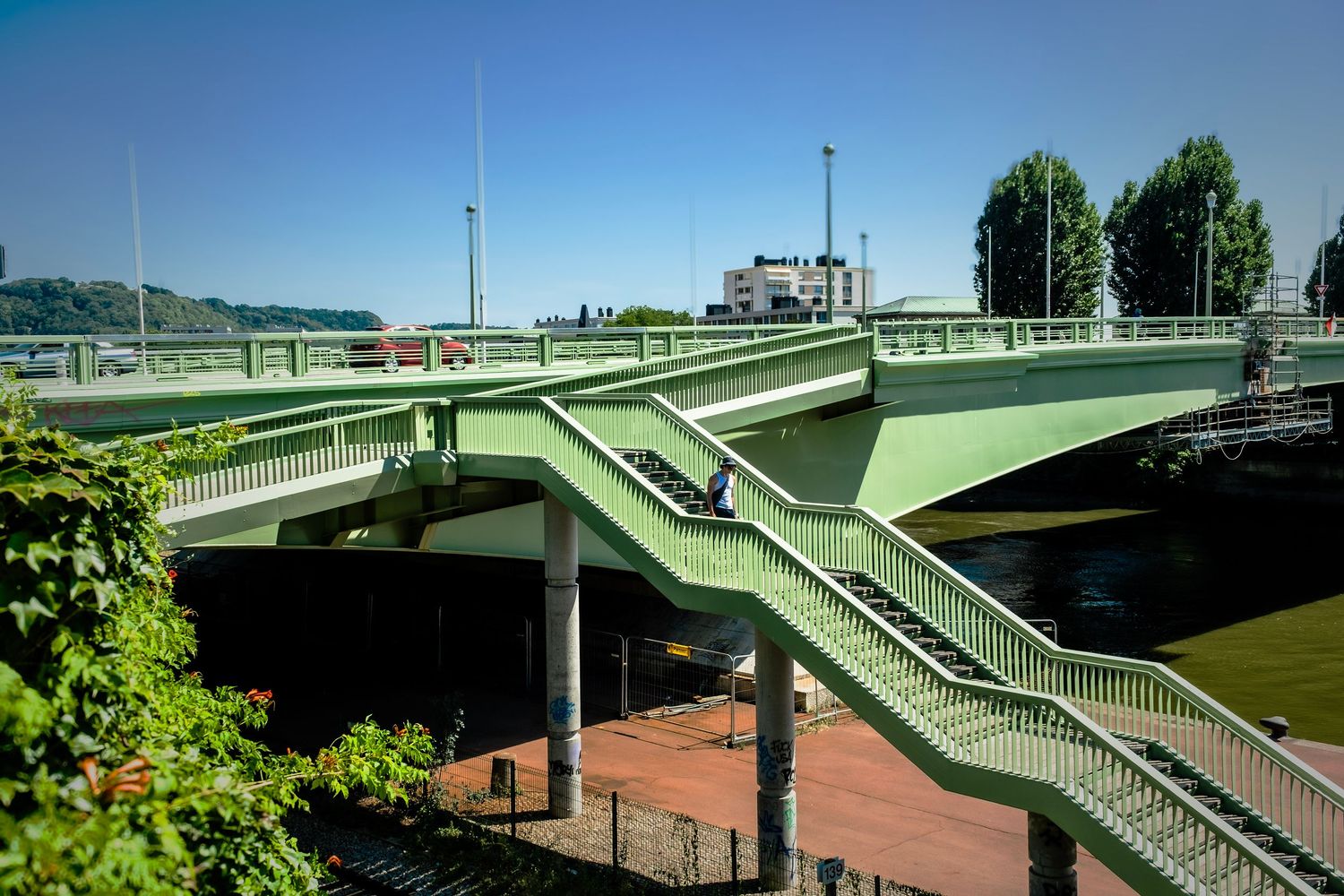Green multi-level bridge with staircases over a river on a sunny day, surrounded by lush trees and brick plaza below.