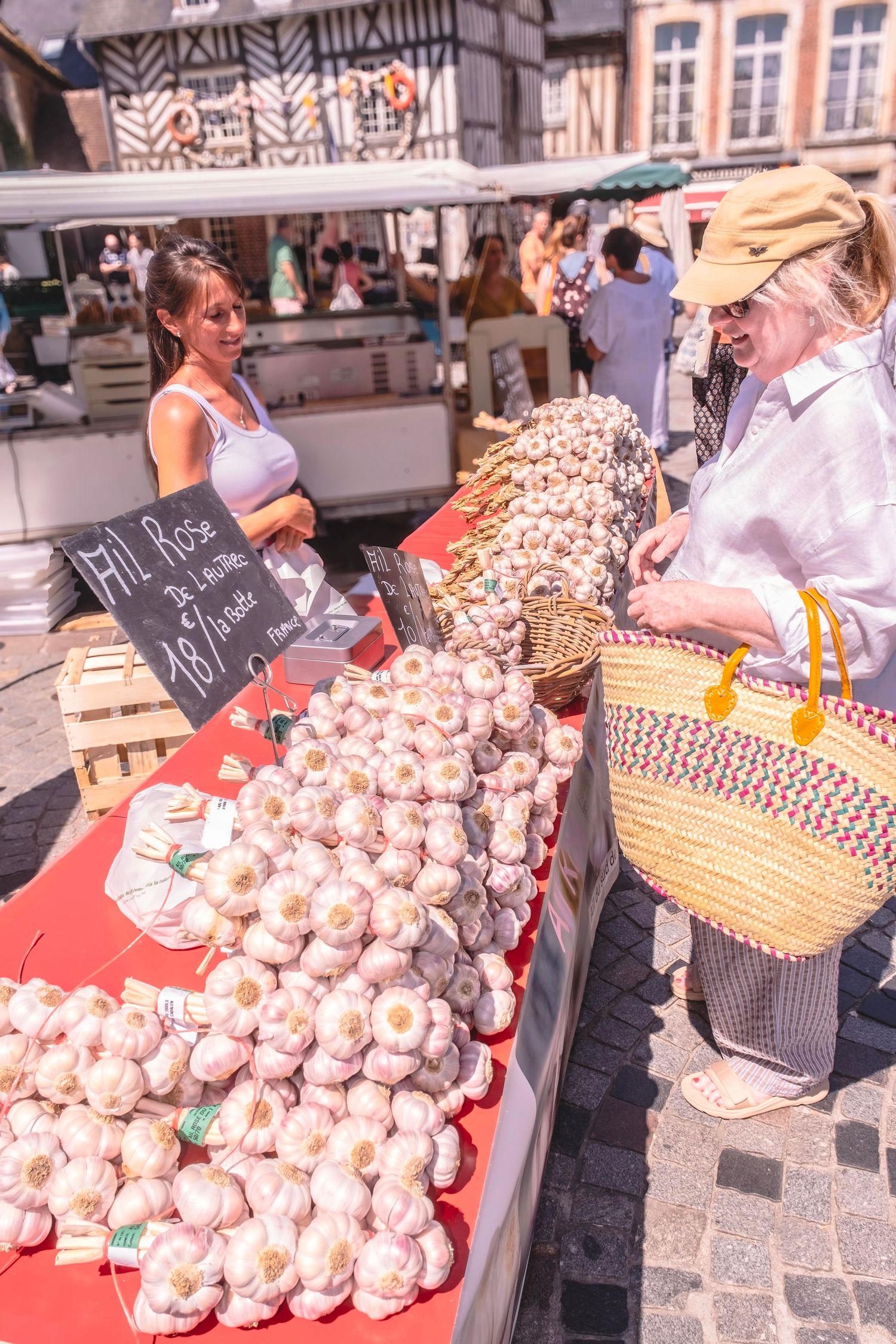 Woman shopping for garlic at a French outdoor market stall with a vendor smiling in the background.