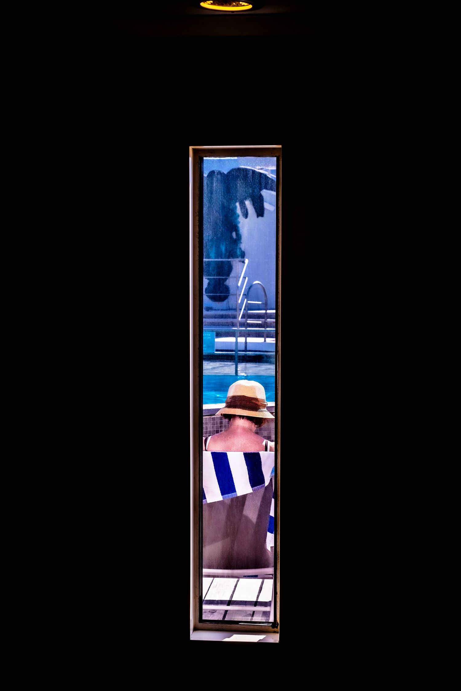 Woman in straw hat and striped towel viewed through narrow window near cruise ship pool ladder.