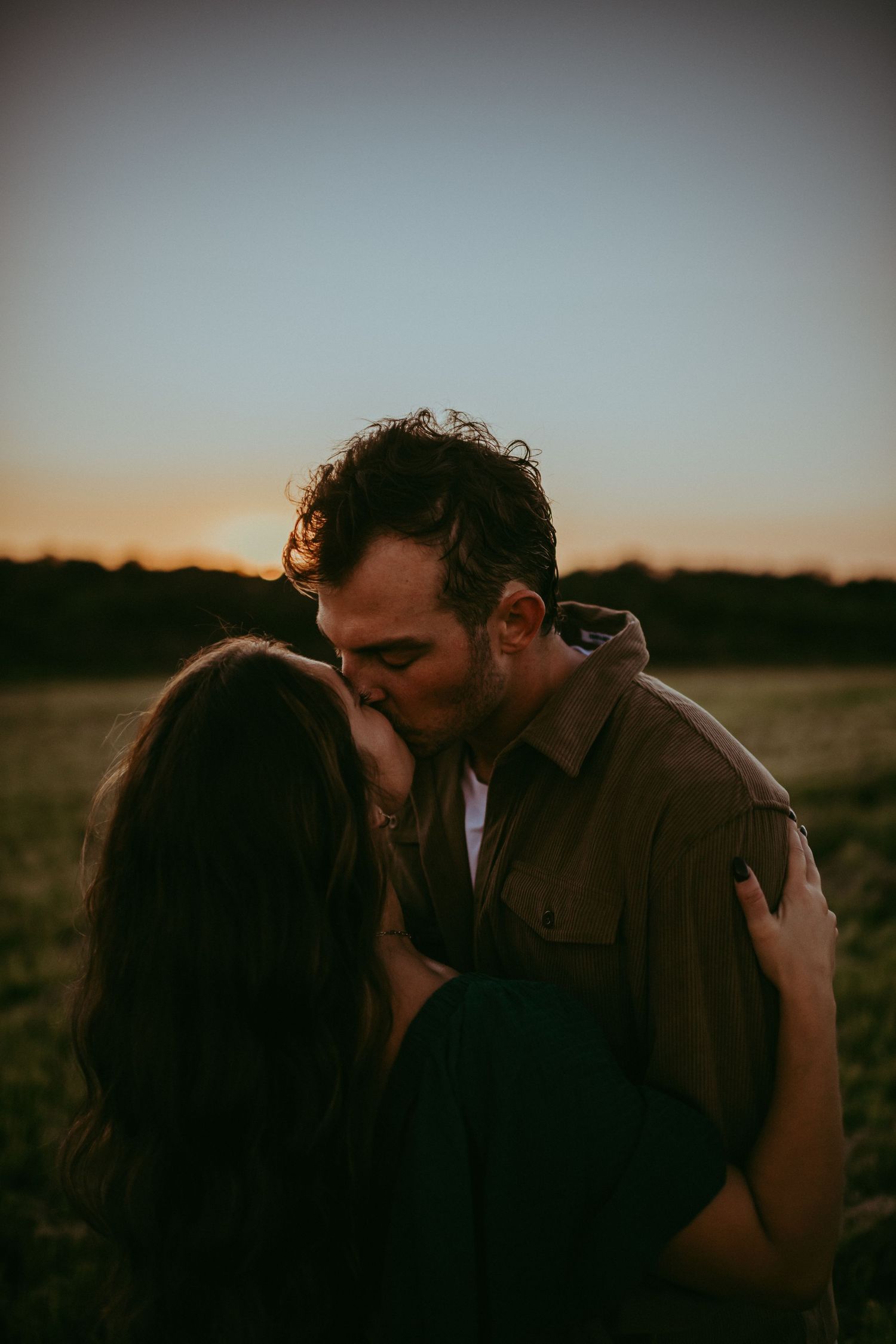 Couple sharing a romantic kiss at sunset in an open field, golden hour light casting a warm glow.