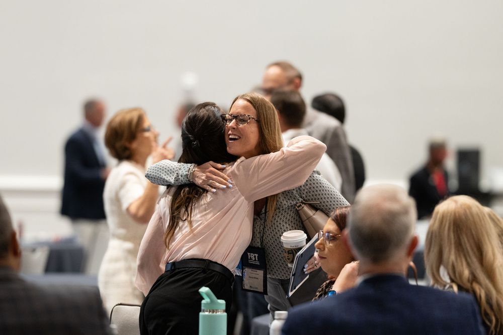 Two women embracing at a professional networking event in a bright conference room with attendees in the background.