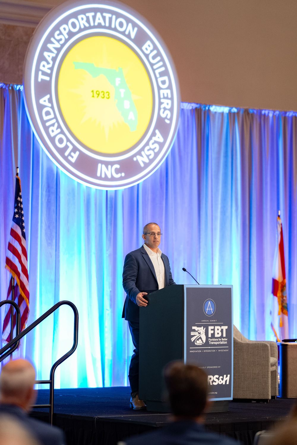 Speaker presents at Florida Transportation Builders Association Inc. annual conference podium with blue stage backdrop.