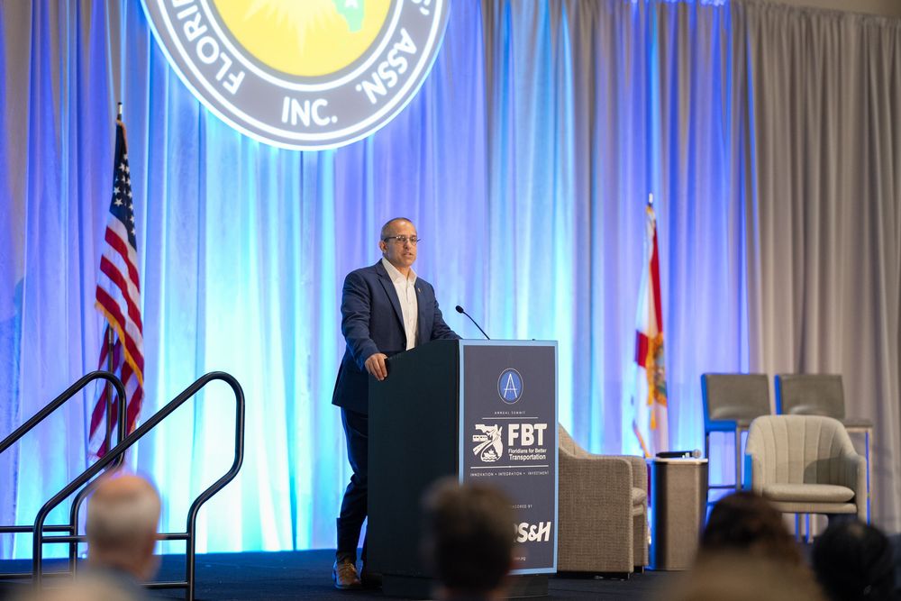 Speaker presenting at FBT conference podium with American and Florida state flags on stage.